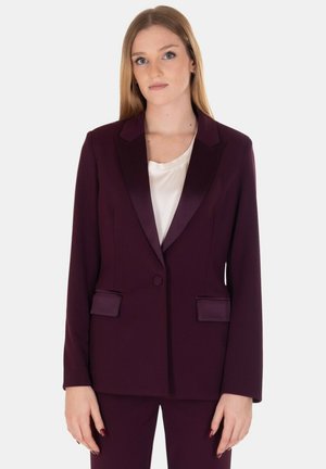 Blazer bordeaux con risvolti in satin, chiusura a un bottone, due tasche frontali, vestibilità strutturata e texture liscia, abbinato a un top crema.