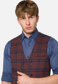 Brązowa kamizelka w kratę wykonana z teksturowanej tkaniny z głębokim dekoltem w kształcie litery V, zestawiona z niebieską koszulą zapinaną na guziki z podwiniętymi rękawami.