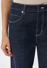 Ciemnoniebieskie dżinsy z białym przeszyciem, przednią kieszenią i zapięciem na guzik. Tkanina wydaje się gładka z delikatną fakturą.