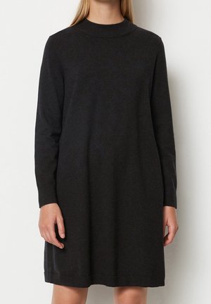 Vrouw gekleed in een effen, zwarte, lange mouws, knie-lange gebreide jurk, staand tegen een effen witte achtergrond.