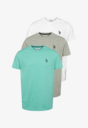 Férfi áll, türkiz rövidujjú pólót visel, kis logóval, sötét navy kék nadrággal és fehér sportcipővel, egyszínű világos háttér előtt.