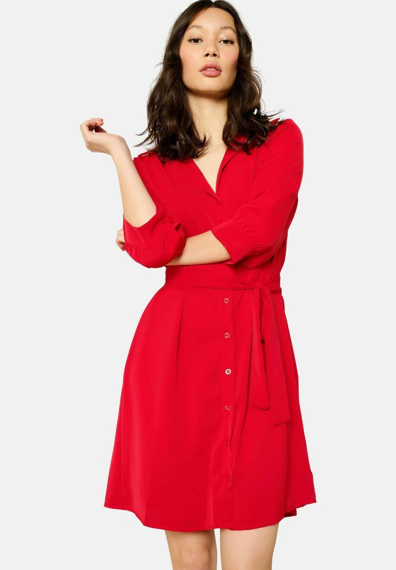 Abito rosso realizzato in tessuto leggero, con davanti a bottoni, colletto e vita con cintura. Maniche lunghe con risvolti e forma a campana.