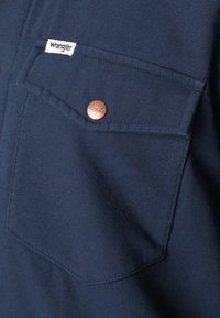 Navy blue fabric shirt featuring a chest pocket with a copper button and Wrangler logo tag. The texture is slightly coarse.