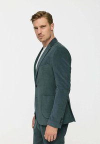 Törökzöld kordbársony blézer, kiszélesedő hajtókával, két elülső zsebbel és egyetlen fekete gombbal záródik, függőleges bordás textúrával.