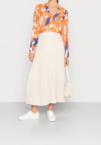Oranžovo-modrý vzorovaný blůza s výstřihem do V a dlouhými rukávy, kombinovaná s plisovanou béžovou maxi sukni. Nosí se s bílými teniskami a světlou taškou.