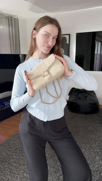 Beige pelzige Handtasche mit überlappendem Klappdeckel, goldfarbenen Beschlägen und einem geflochtenen Kettenriemen, gehalten vor einer Person, die ein hellblaues Oberteil trägt.