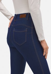 Jeans azul oscuro con costuras naranjas, de cintura media y dos bolsillos traseros. Elaborados con denim de textura suave y un parche de logo de cuero.