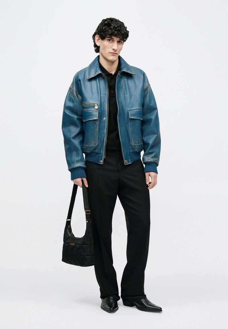 Young man with curly hair wears blue leather jacket, black pants and shoes, holding a black quilted handbag against a plain background.