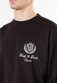 T-shirt en coton noir avec un col rond, logo brodé avec détail de couronne, et texte "Famille & Amis" et "Pegador".