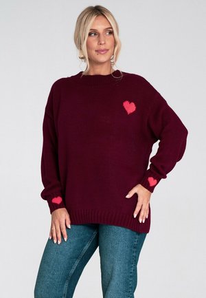 Burgundowy sweter dzianinowy o luźnym kroju, z różowymi akcentami serca na klatce piersiowej i rękawach, z prążkowanymi mankietami i dolnym brzegiem. Połączony z dżinsami.