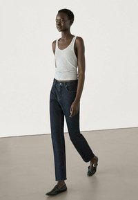 Femme marchant portant un haut sans manches gris, un jean droit foncé et des chaussures noires à boucle sur un fond clair uni.