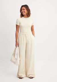 Krēmkrāsas īsām piedurknēm tops, kas savienots ar bēšiem platām biksēm, kurām ir elastīga josta un sānu kabatas, papildināts ar baltu rokassomu.