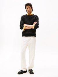 Μαύρο πλεκτό πουλόβερ με σχέδιο καλίγραμμο πάνω από ένα κίτρινο πουκάμισο με κολάρο, συνδυασμένο με λευκά παν trousers και μαύρες loafer, σε ποζάρισμα μπροστά από λευκό φόντο.