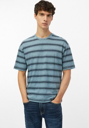 Jonge man met krullend haar, gekleed in een blauw gestreept T-shirt met korte mouwen en donkere jeans, handen in de zakken, staand tegen een effen achtergrond.
