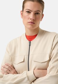 Béžová bomber bunda se dvěma předními kapsami, žebrovaným límcem a manžetami a zipem na zapínání. Texturovaná látka, minimalistický design, decentní značení.