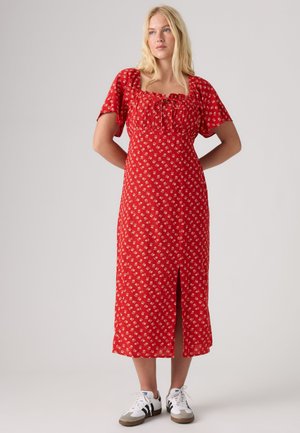 Frau steht in einem roten, geblümten Midi-Kleid mit Puffärmeln und vorderem Schlitz, trägt weiße Sneakers, vor neutralem Hintergrund.