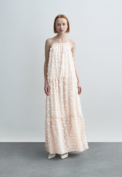 Robe longue en tissu rose doux, ornée d'appliqués floraux, design à étages et fines bretelles. Texture lisse avec une silhouette fluide.