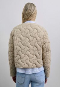 Chaqueta acolchada beige con un patrón ondulado, de corte amplio y un diseño ligeramente recortado, llevada sobre una camisa azul clara.
