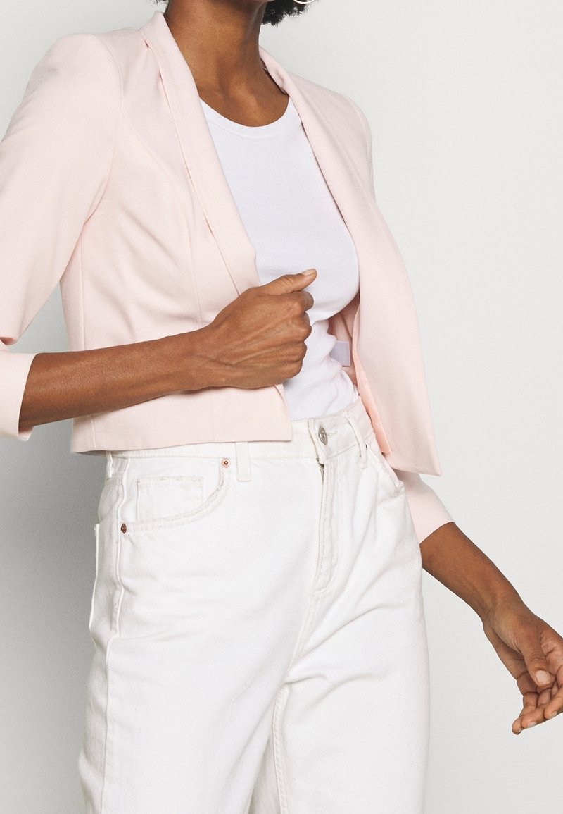 Vrouw draagt een lichtroze cropped blazer over een wit T-shirt en witte high-waisted jeans, en past de blazer aan met één hand.