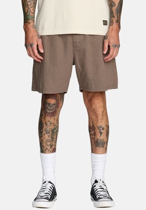 Bruine shorts met een losse pasvorm, gemaakt van een gestructureerde stof. Draag ze met witte sokken en zwarte canvas sneakers. Gedetailleerde tatoeages zichtbaar op de benen.