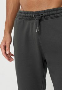 Graue Sweatpants mit elastischem Bund und Kordelzug. Sie haben eine glatte Textur und eine Seitennaht, mit einer Nahaufnahme des Bundes und einer Hand.