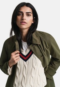 Trench coat verde oliva sobre un suéter de punto con trenzas color crema y escote en forma de V azul marino y rojo. Cuenta con un cierre de botón y un ajuste relajado.