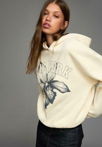 Młoda kobieta z długimi brązowymi włosami, ubrana w kremowy hoodie z napisem "Nowy Jork" i dużym czarnym kwiatowym wzorem, pozująca na szarym tle.