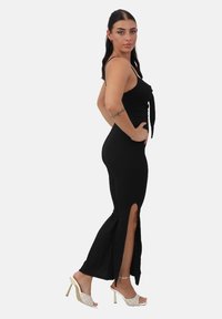 Robe noire côtelée avec une silhouette ajustée, des bretelles spaghetti et des fentes sur les côtés. Le mannequin porte des talons blancs avec un motif matelassé.