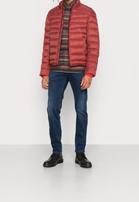 Rød oppustelig jakke med høj krave, kviltet design, parret med en ternet skjorte og mørkeblå jeans. Sorte snørebåndsstøvler fuldender looket.