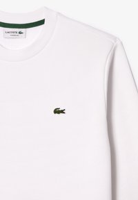 Valkoinen Lacoste klassinen sovitus huppari, jossa on vihreä krokotiili-logo brodeerattuna vasemmalla rintamuksessa ja vihreä kauluksen yksityiskohta kauluksen sisäpuolella.