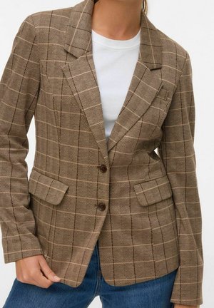 Femme portant un blazer marron à carreaux à deux boutons sur une chemise blanche et un jean bleu, main tenant l'ourlet du blazer.
