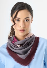 Mehrfarbiger Schal mit grauem Grund, burgunderfarbener Umrandung und bedruckten Designs sowie Fransen. Umschlungen um den Hals in einer dreieckigen Form.