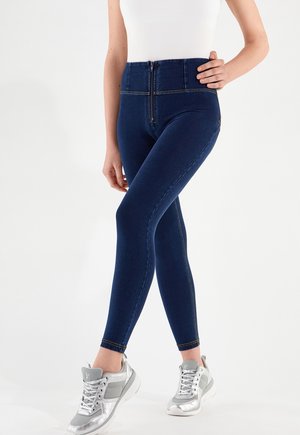 Nainen pukeutune korkeavyötäröisiin, tummansinisiin skinny-farkkuihin, joissa etutaskun vetoketju ja hopeanväriset urheilukengät, seisoo yhdellä kädellä lanteilla.
