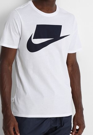 Homme portant un t-shirt blanc à manches courtes avec un grand motif abstrait noir sur la poitrine et un pantalon sombre.
