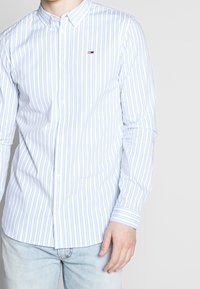 Camicia button-up a righe in azzurro chiaro e bianco, con righe verticali, colletto e un piccolo logo sul petto. Tessuto in cotone.