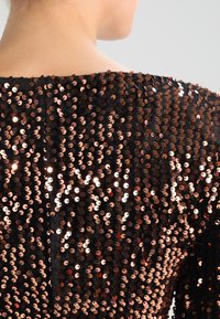 Svart topp täckt med rosaguldiga paljetter som visar en texturerad, glittrande yta. Halsringningen är vid, vilket avslöjar ett slätt tyg under.