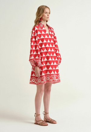 Junge Frau steht und trägt ein rot-weiß gemustertes Kleid mit geometrischem Muster, Puffärmeln und beigen flachen Sandalen vor einem schlichten Hintergrund.