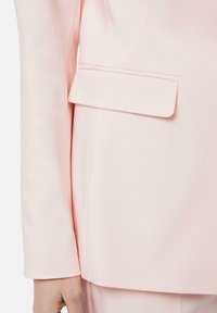 Manica di un blazer rosa con un design elegante, dotato di una singola tasca a patta. Il tessuto ha una texture liscia e una leggera lucentezza.