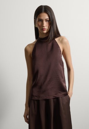 Mujer con cabello lacio y oscuro, vestida con un top de satén marrón sin mangas y una falda marrón, de pie frente a un fondo claro y liso.