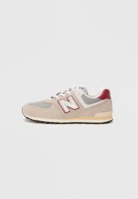 Basket de New Balance beige et grise avec des lacets blancs, des accents rouges et une semelle grise tachetée de noir, vue de côté sur un fond blanc.