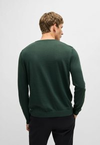 Maglione lavorato a maglia verde scuro con scollatura rotonda, maniche lunghe e una finitura leggermente testurizzata. Presenta dettagli sulle cuciture laterali all'orlo.