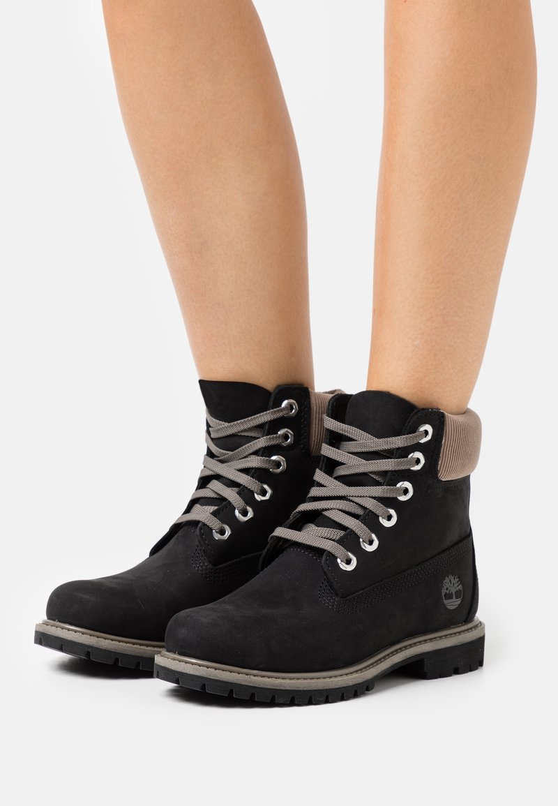 Bottines en daim noir avec lacets gris, col rembourré et semelle en caoutchouc. Caractéristiques œillets argentés et logo sur le côté.