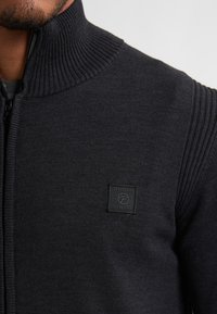 Schwarzer Zip-Up-Pullover mit geripptem Kragen und Schulterdetails; verfügt über ein quadratisches Logo-Label auf der Brust aus kontrastierendem Material.