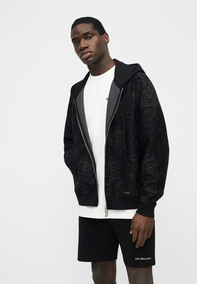 Bărbat tânăr purtând un hanorac negru cu fermoar și model, tricou alb și pantaloni scurți negri cu textul „LES BENJAMINS” pe piciorul stâng.