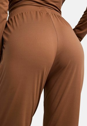 Nahaufnahme einer Person, die braune High-Waist-Hosen mit gerafftem elastischem Bund und ein langärmliges Oberteil trägt, stehend vor einem schlichten Hintergrund.