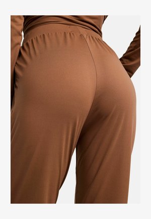 Nahaufnahme einer Person, die braune High-Waist-Hosen mit gerafftem elastischem Bund und ein langärmliges Oberteil trägt, stehend vor einem schlichten Hintergrund.