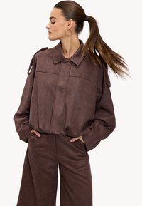 Femme avec une longue queue de cheval portant une veste oversized marron foncé et un pantalon large assorti, les mains dans les poches, faisant face à gauche les yeux fermés.