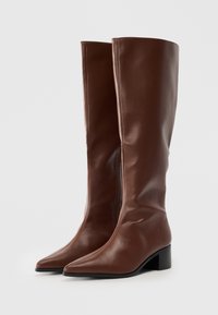 Une paire de bottes en cuir marron hautes avec des pointes effilées et de petits talons carrés, placées côte à côte sur une surface blanche.