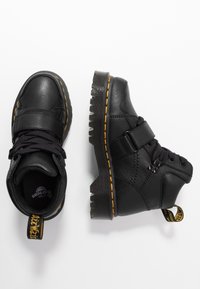 Černé kožené polobotky s tkaničkami a suchým zipem, které mají žluté prošívání a strukturovanou gumovou podrážku s drážkami.