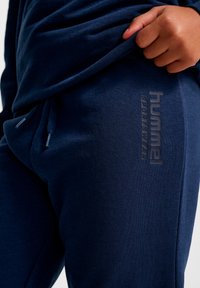 Granatowe spodnie dresowe wykonane z miękkiego, teksturowanego materiału. Posiadają elastyczną talię ze sznurkami oraz monochromatyczne logo po boku.